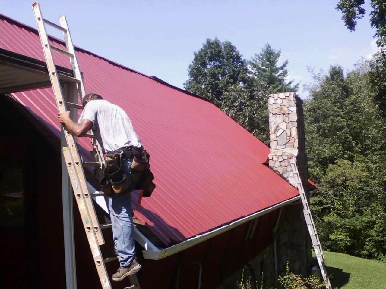 Roofer on a ladder beside a red metal roof and stone chimney — Soffit & Fascia Repair by Roofing Brothers in Wanaque